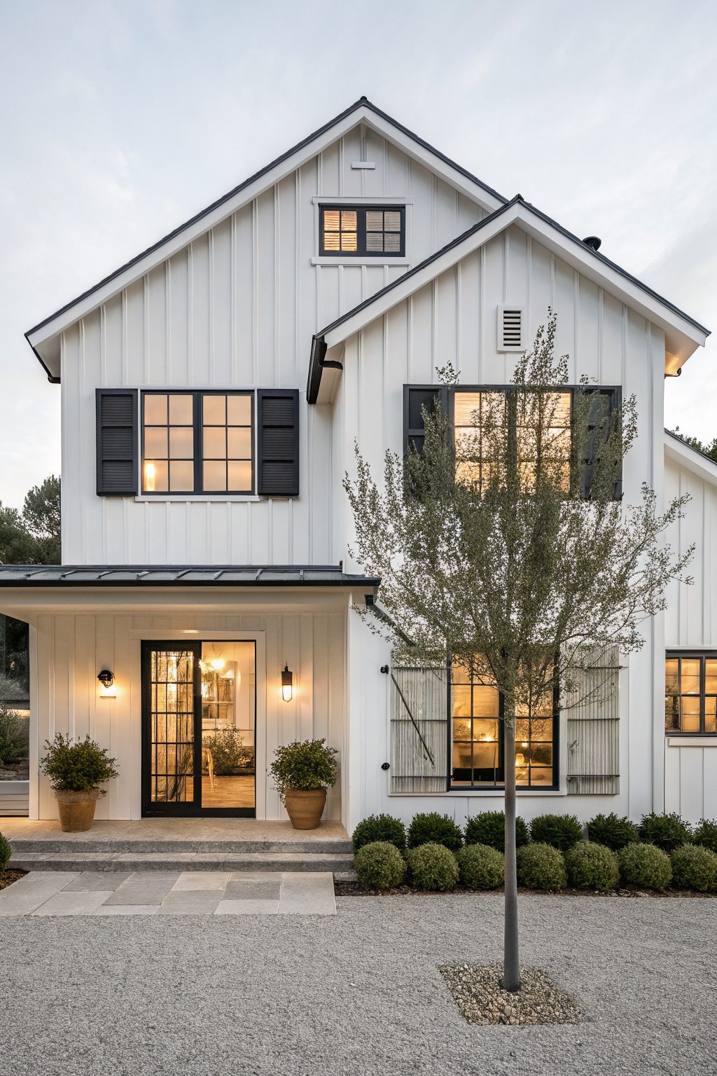 A two-story white farmhouse with black window frames, black shutters, and a black front door, shown from the front with a small tree and gravel driveway.