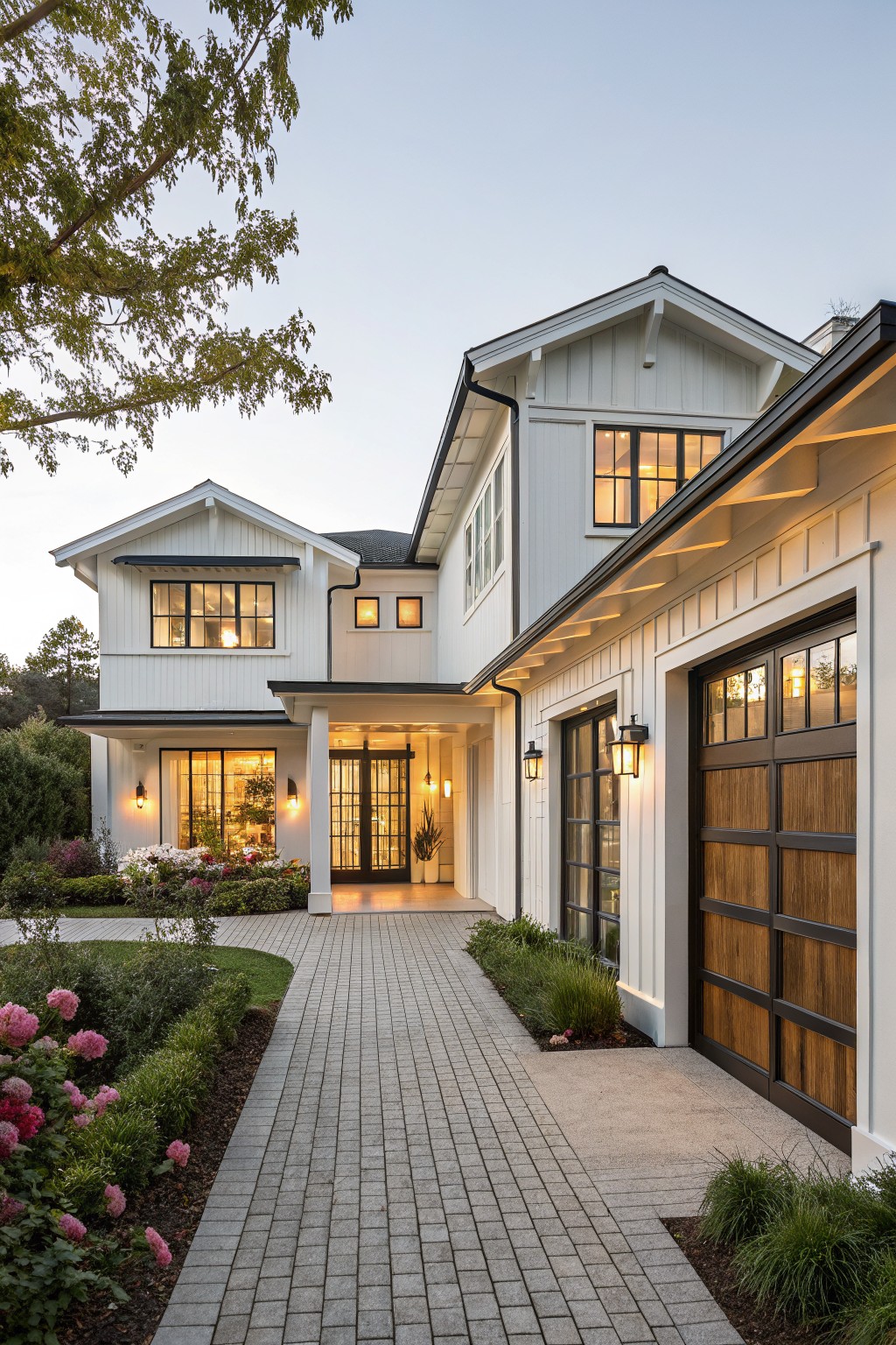 Two-story white farmhouse with lit windows, brick driveway, and wooden garage at dusk