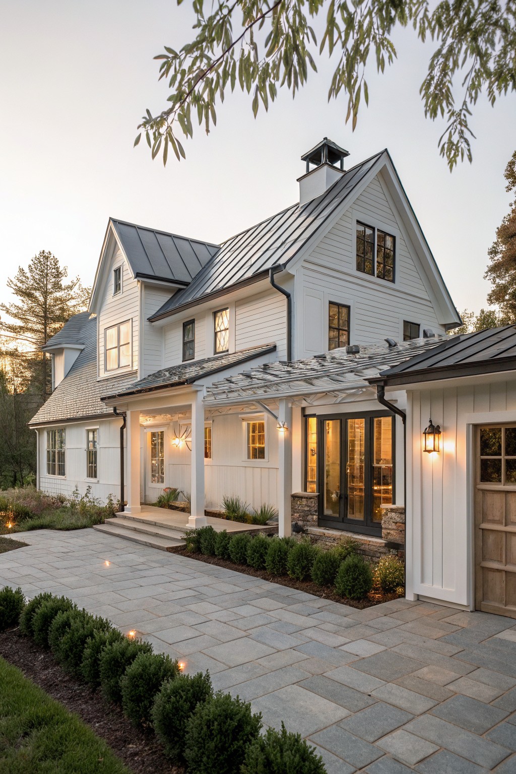 A white two-story farmhouse with dark metal roofs, black window frames, stone accents, and a paved driveway bordered by shrubs.