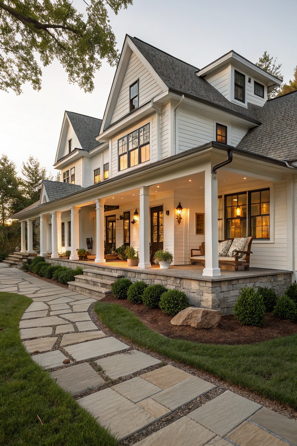 A white two-story farmhouse with a wide wraparound porch supported by white columns, stone foundation, black-framed windows, and a stone pathway in the foreground.