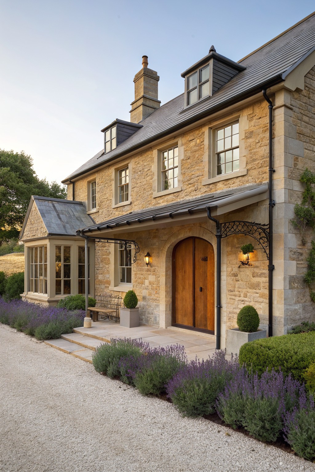A two-story stone farmhouse with an arched wooden front door, multiple multi-pane windows, a dark slate roof, and lavender plantings along the gravel driveway.