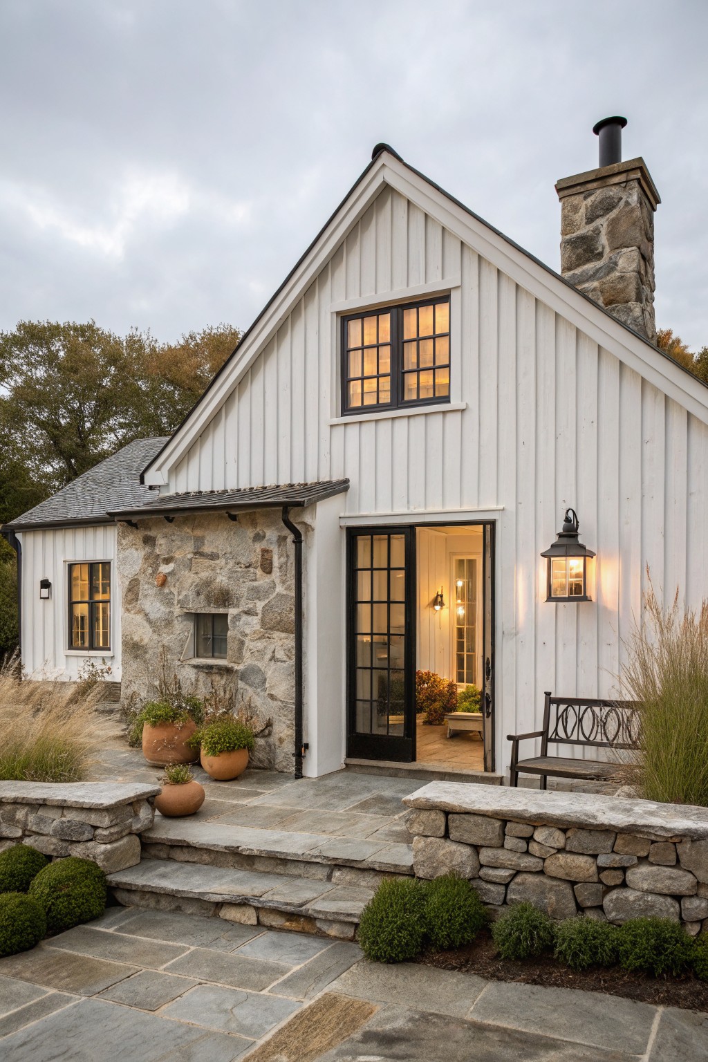 A white vertical siding farmhouse with a stone chimney and partial stone wall at the entry, black-framed door and windows, and stone steps leading to the front.
