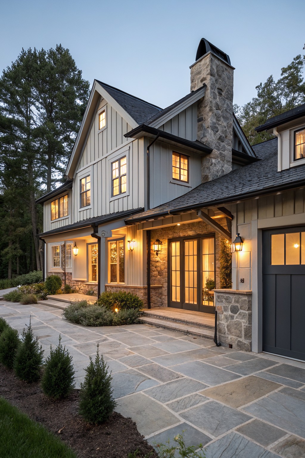 Two-story gray house with stone chimney, lit windows, and paved courtyard at dusk