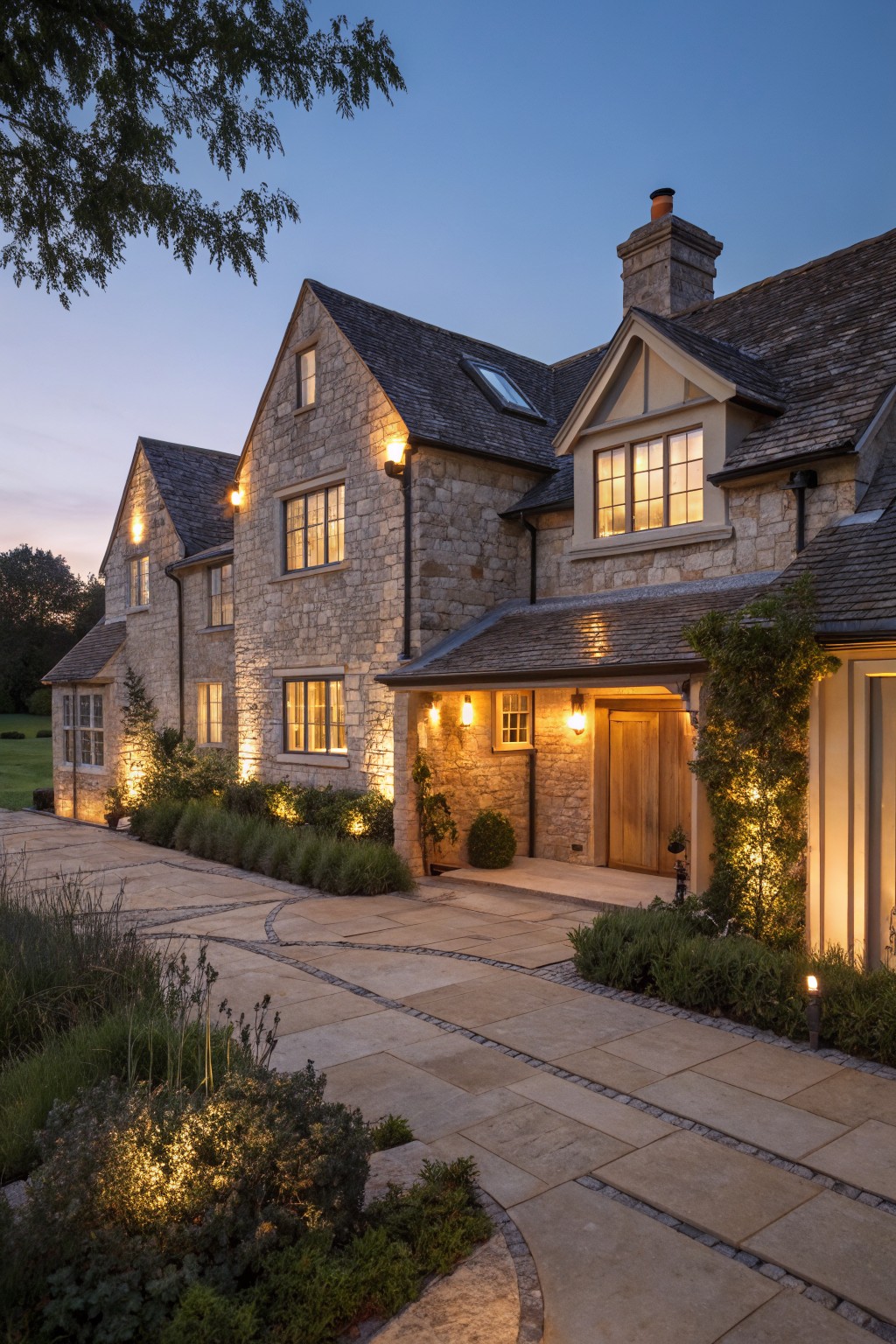 A large stone farmhouse with multiple gables, a wooden front door, and warm exterior lighting along the driveway at dusk.