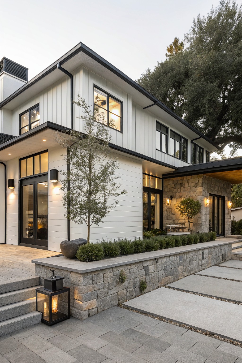 Modern farmhouse exterior with white vertical siding, black trim, stone accents on the lower walls, and a matching stone retaining wall with planting beds in front.