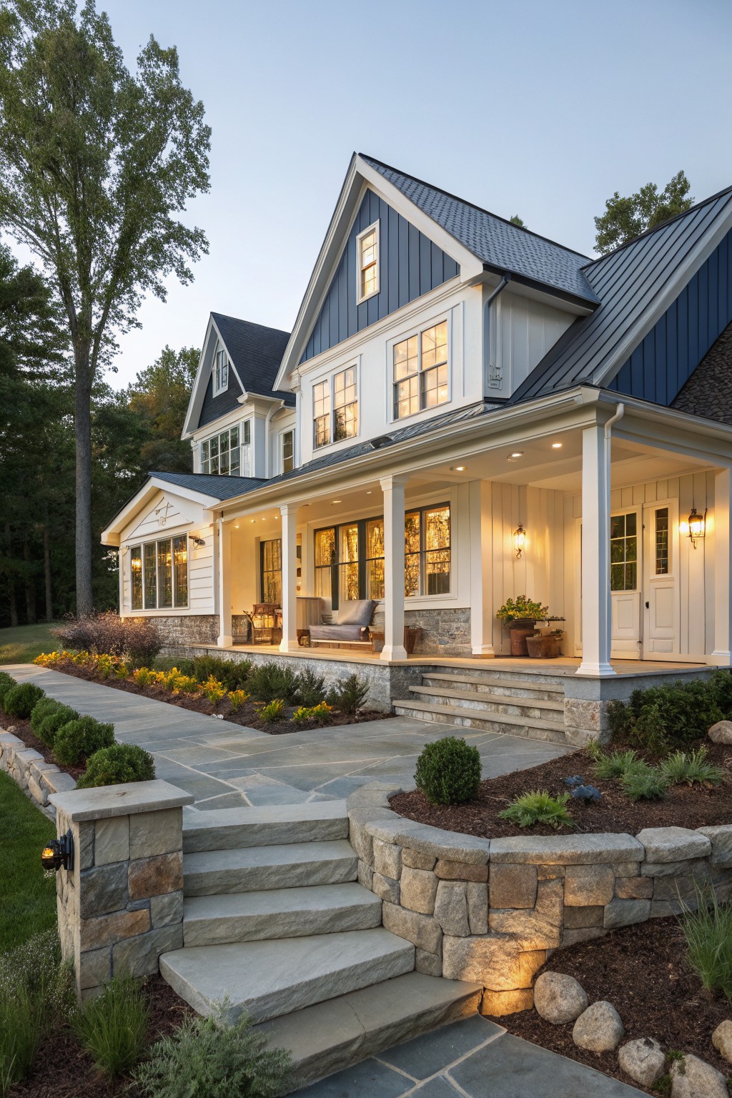 A two-story traditional farmhouse with white siding, dark blue gables, a large covered front porch supported by white columns, and stone steps leading up from a landscaped yard.