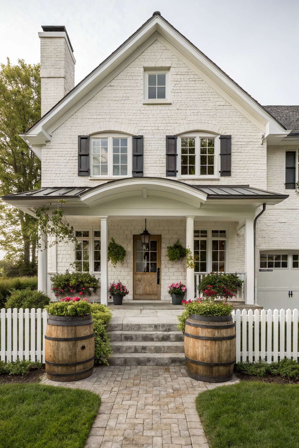 White brick house with porch, steps, barrel planters, and picket fence.