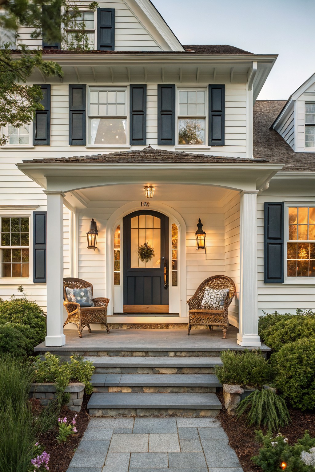A white Cape Cod house with dark shutters and a covered front portico featuring an arched entry, two wicker chairs, lanterns, and stone steps.