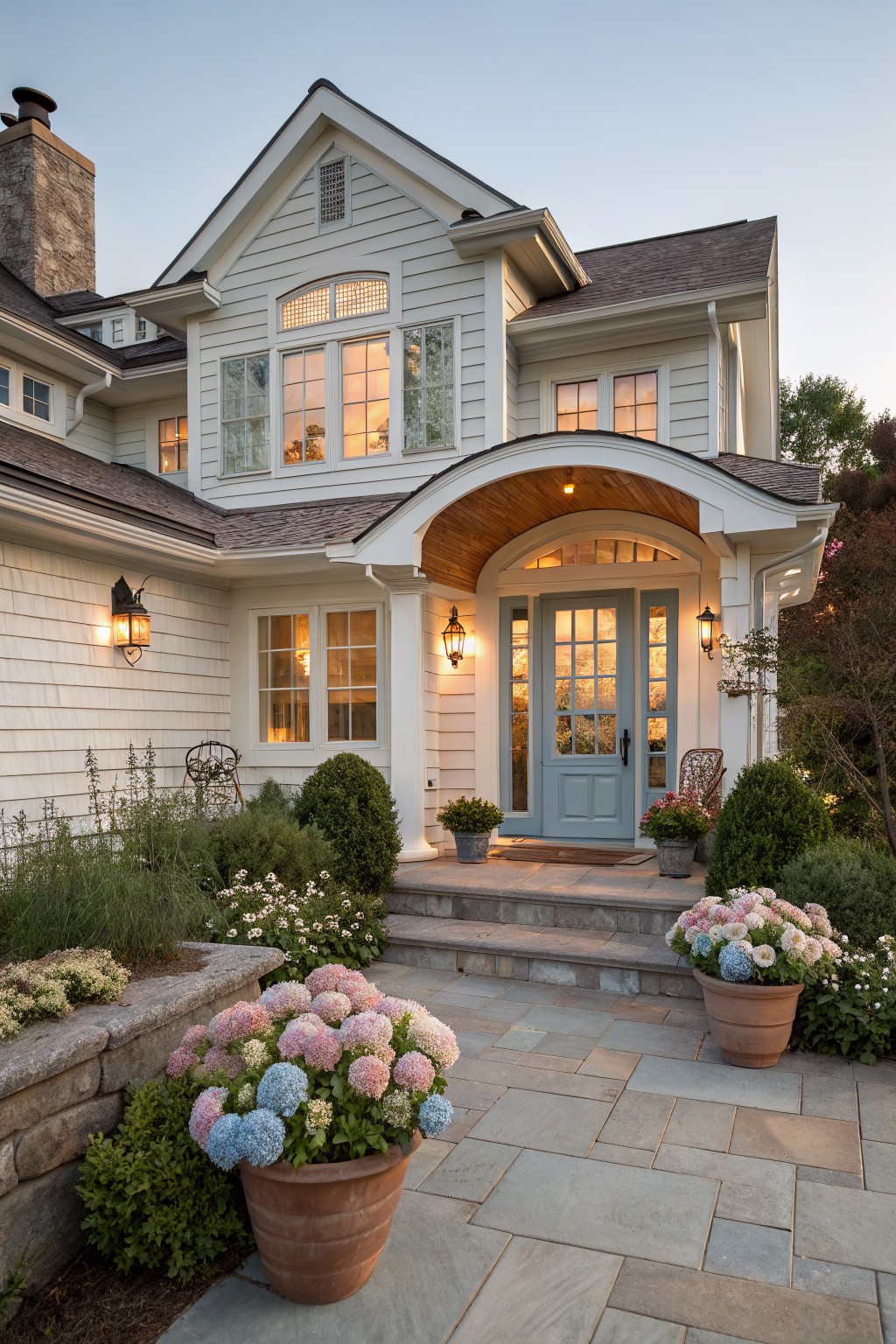 A white two-story Cape Cod farmhouse with a light blue front door under a curved wooden portico, stone steps, and large potted hydrangeas along the walkway.