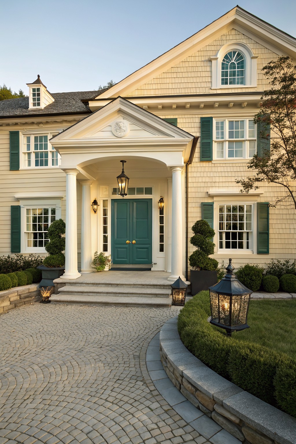 A Cape Cod farmhouse exterior featuring a white portico with columns framing a teal double door, flanked by windows with dark green shutters and stone steps leading to a cobblestone driveway.