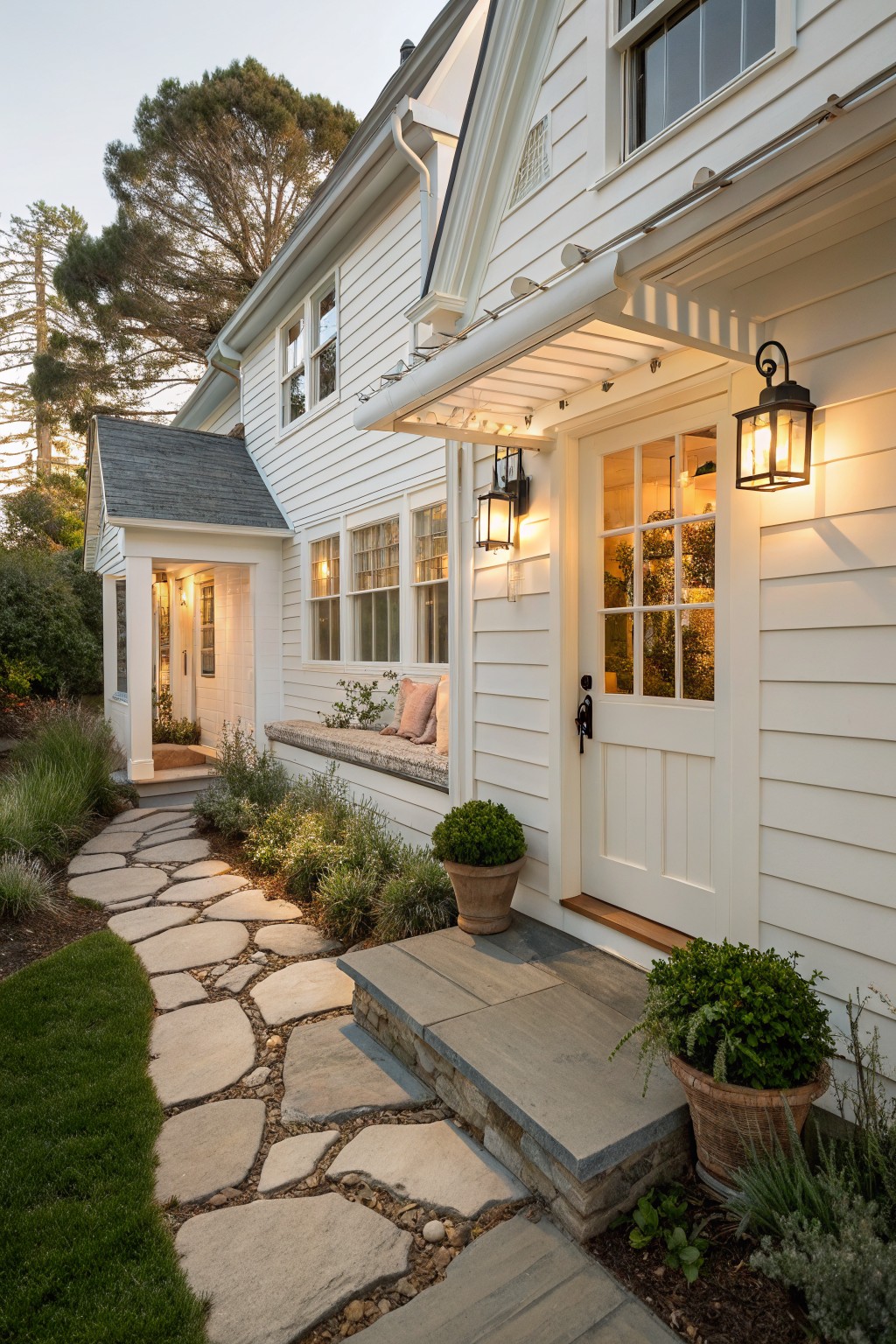 A white Cape Cod farmhouse exterior with a winding stone pathway and steps leading to a white front door, flanked by potted plants and wall lanterns.