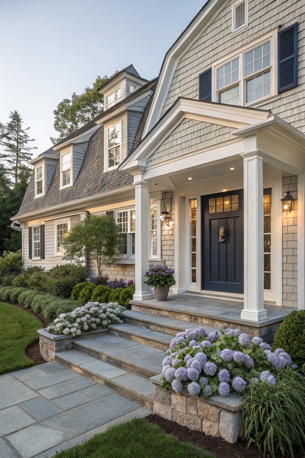 A two-story house with light gray blue siding, white trim, a dark blue front door, and stone steps lined with shrubs.