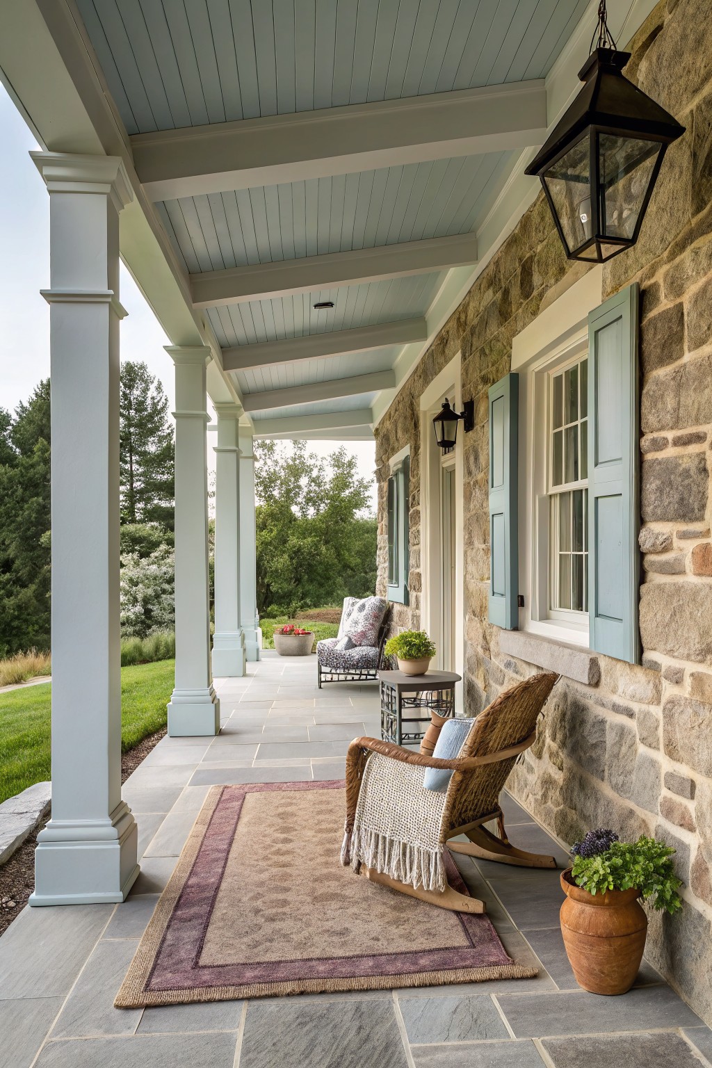 A covered stone porch with white columns, a wooden rocking chair on a large neutral rug, light blue shutters, and potted plants along the wall.