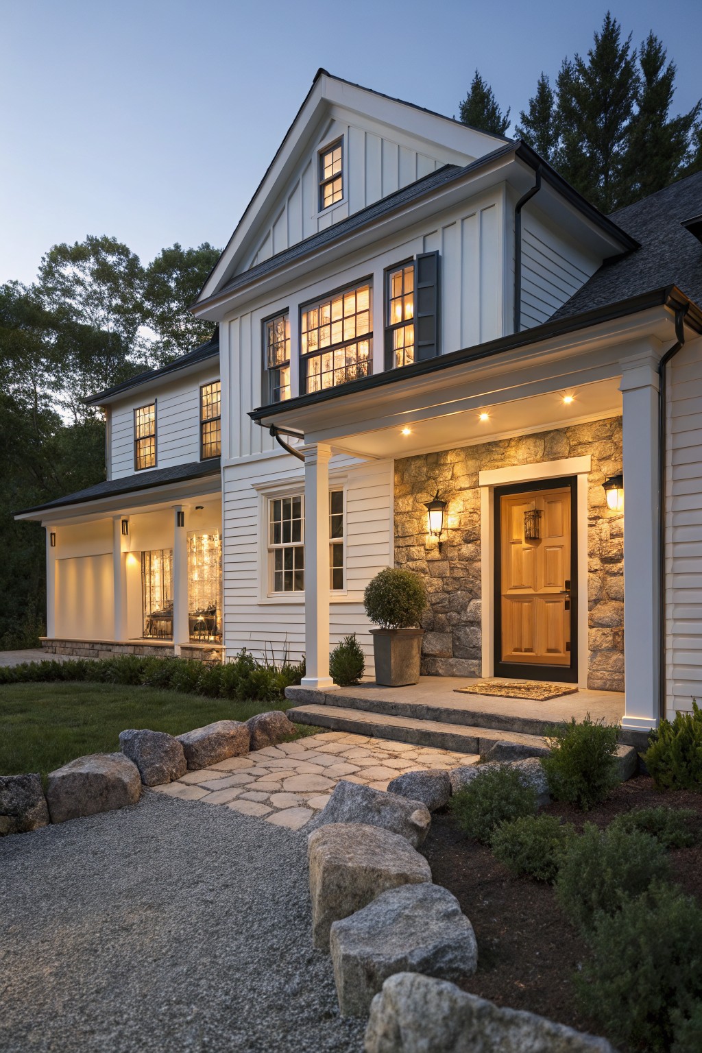 White two-story farmhouse with stone veneer framing a wooden front door, lit by wall sconces at dusk.