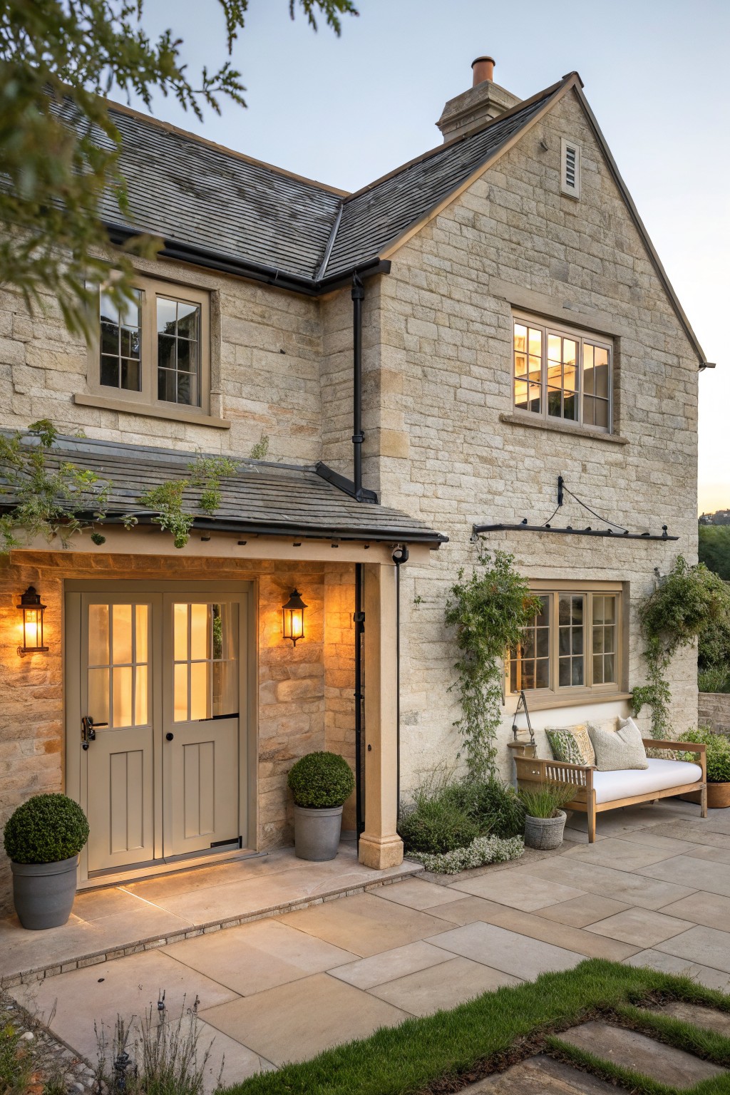 A stone house exterior featuring double front doors in a soft neutral tone, wall lanterns, a paved patio with a wooden bench, and climbing plants along the walls.