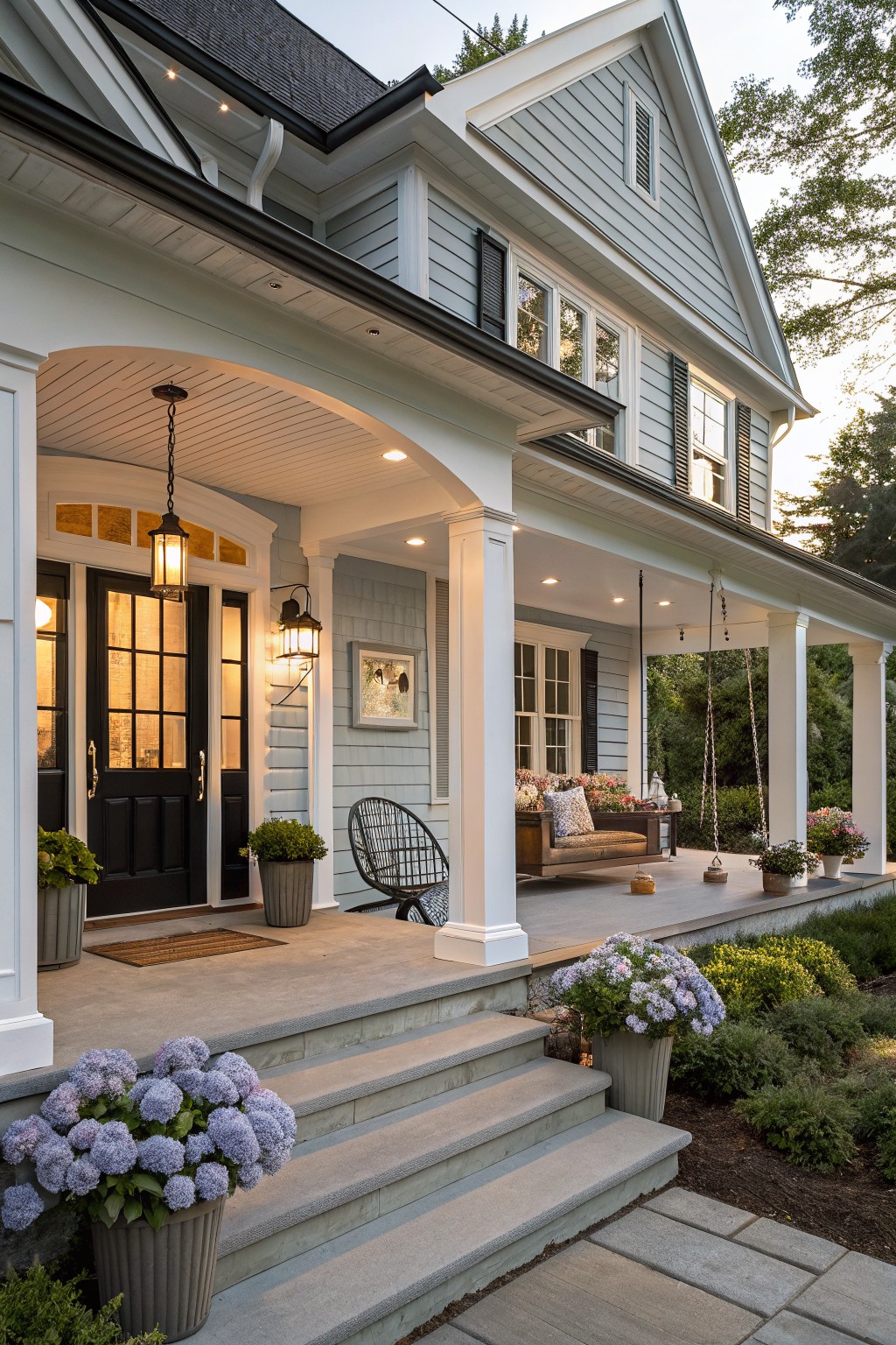 A light blue farmhouse with white trim, a black front door under a white arched porch ceiling, and steps flanked by potted hydrangeas.