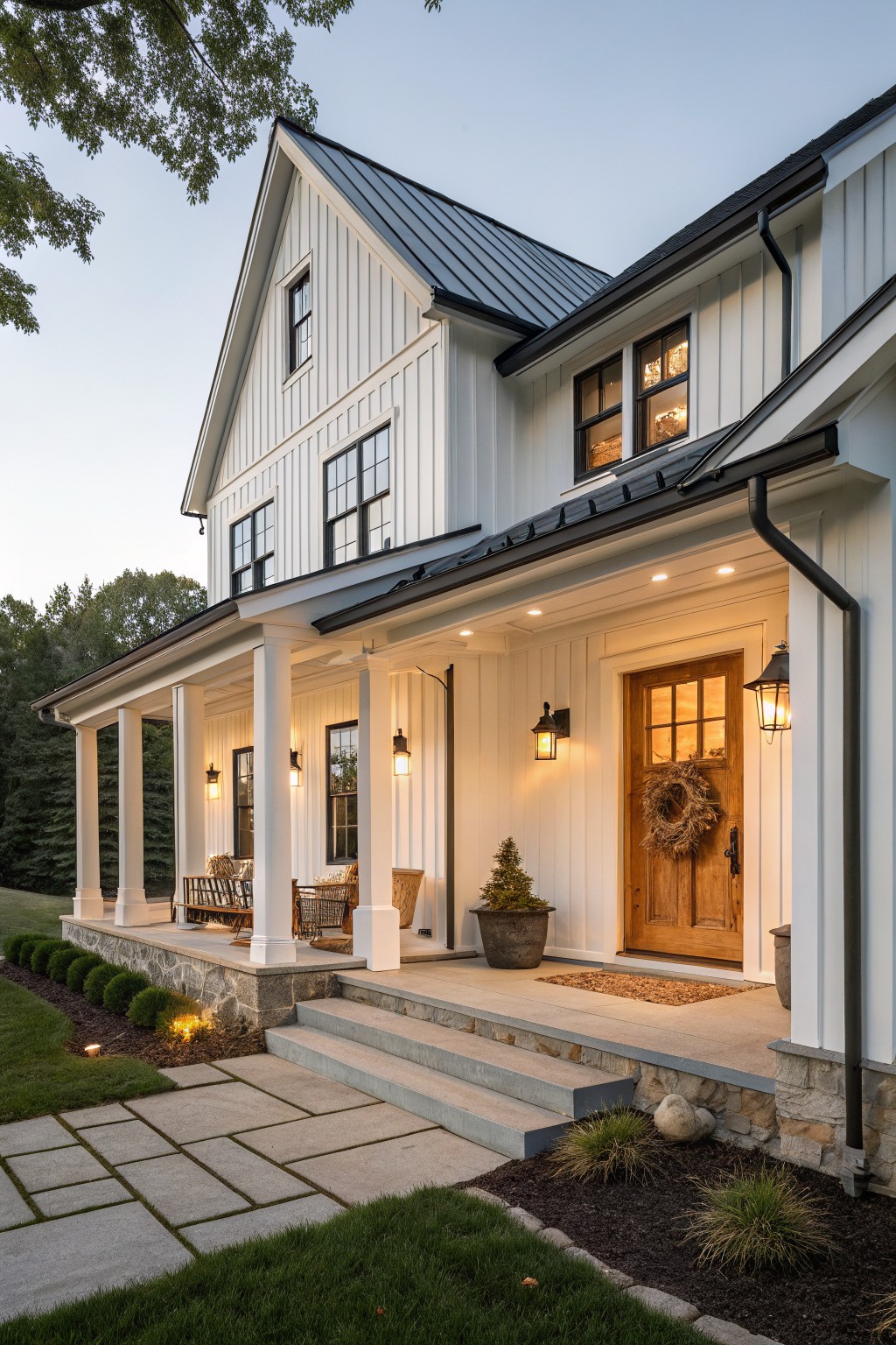 A two-story white farmhouse with vertical siding, black metal roof, covered porch with white columns, stone foundation, and a centered wooden front door with a wreath.