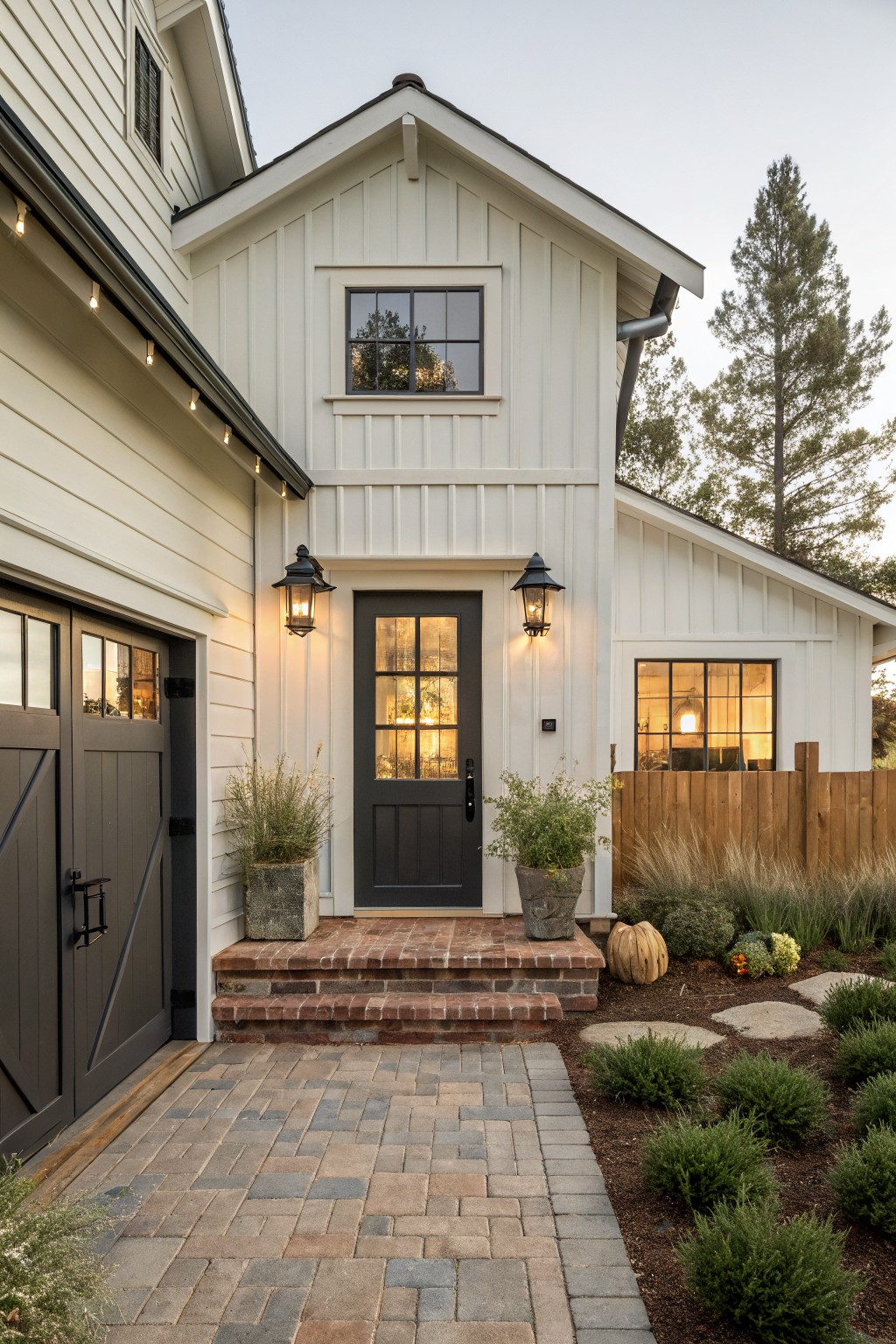 A white farmhouse exterior featuring a dark front door, black lanterns on either side, brick steps, and a paved walkway.