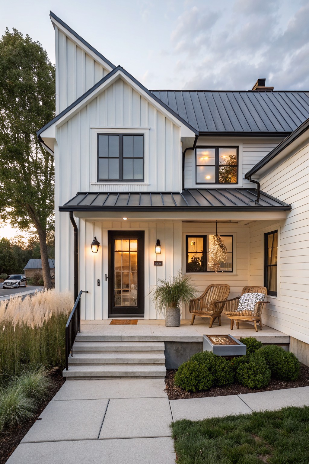 Front view of a white two-story farmhouse with black metal roofing, a black front door, and a covered porch with two wooden chairs.