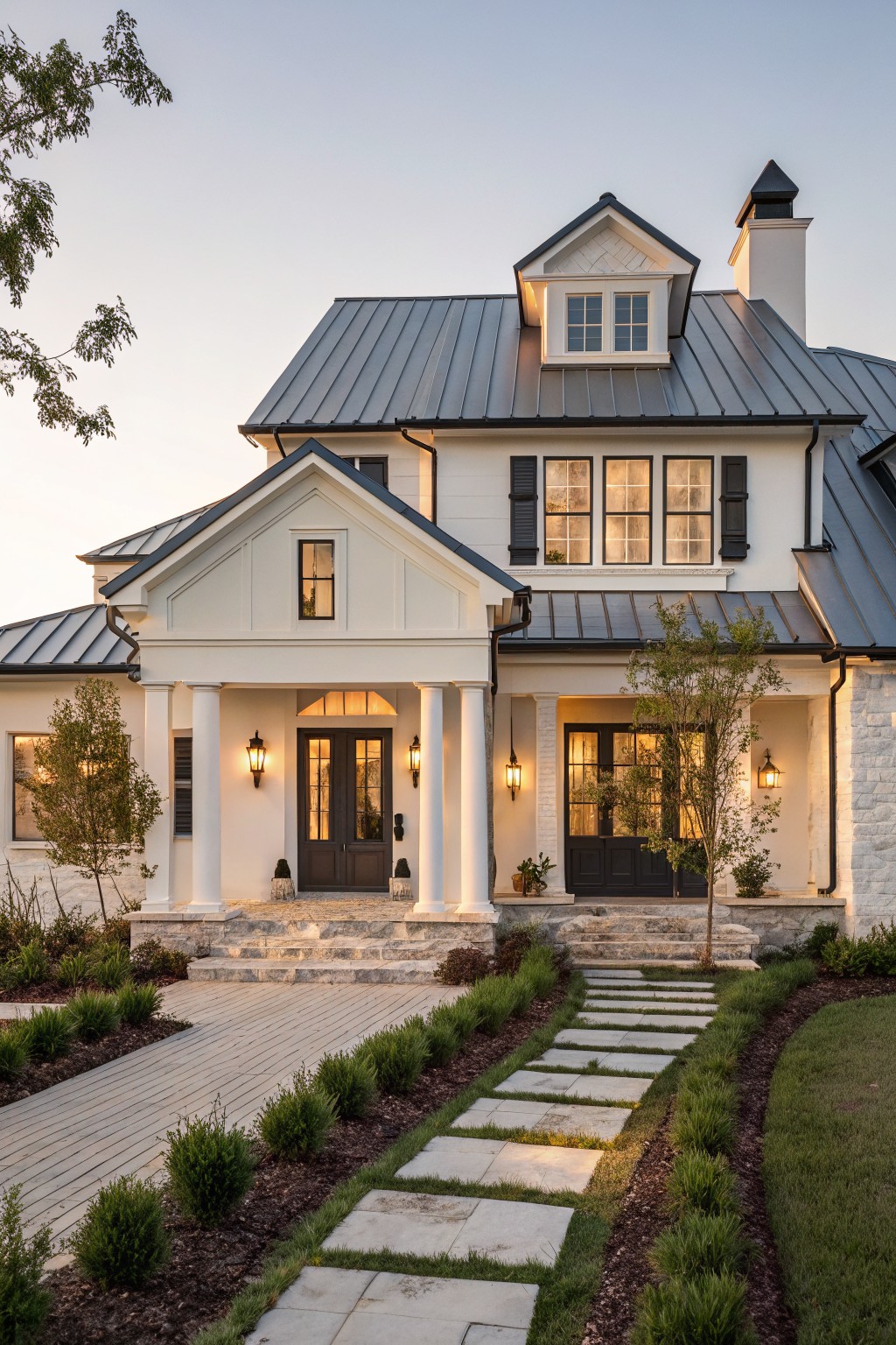 A white farmhouse exterior with a dark metal roof, gabled dormer, front porch with white columns, and stone steps.