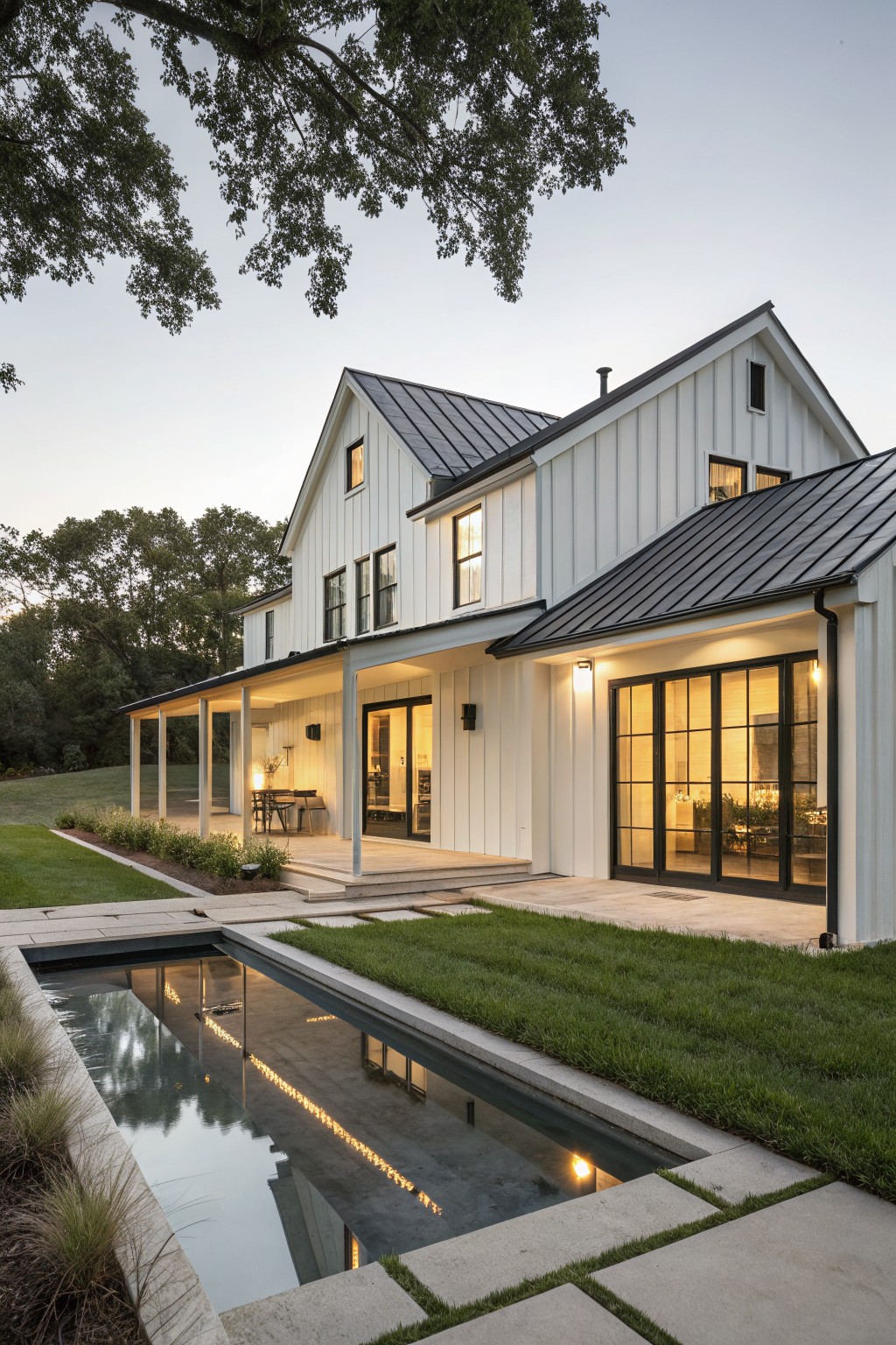 A white two-story farmhouse with a dark metal roof, large glass doors, and a long rectangular pool in the front yard.