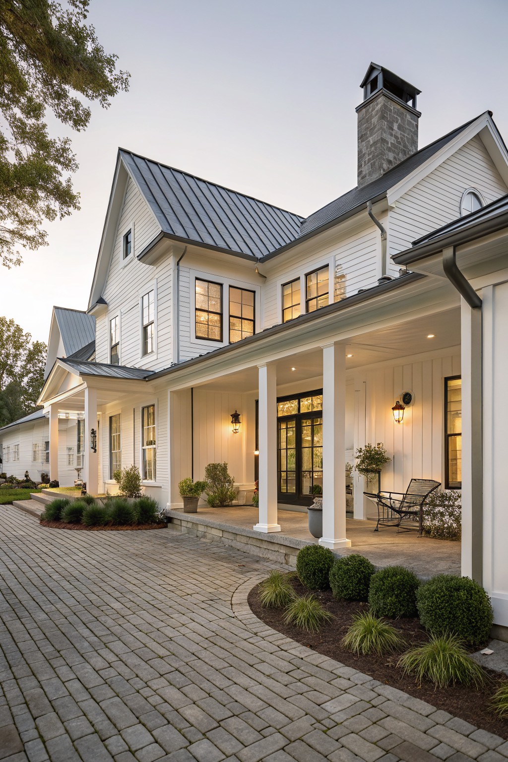 White two-story house with porch, stone chimney, and brick driveway at dusk