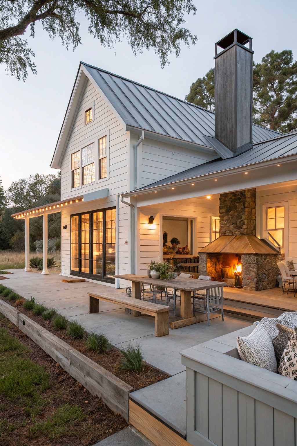 White farmhouse with gray metal roof, black windows, and covered patio with stone fireplace.