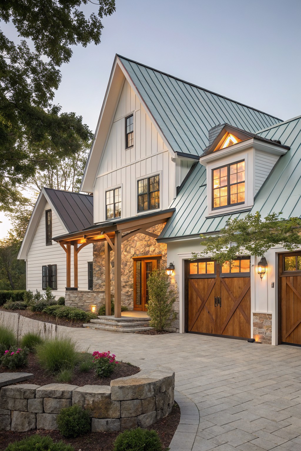 A white vertical siding farmhouse with a green metal roof, stone foundation accents, wooden garage doors, and a paved driveway.