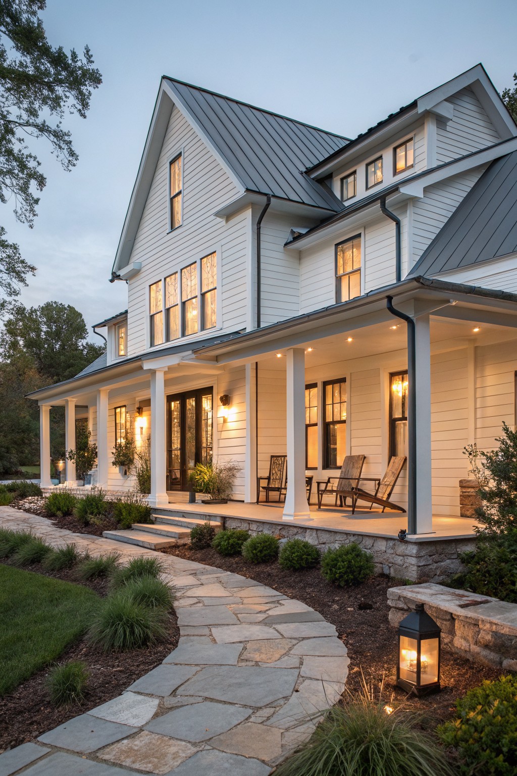 A white two-story farmhouse with a dark metal roof, front porch, and stone foundation at dusk.
