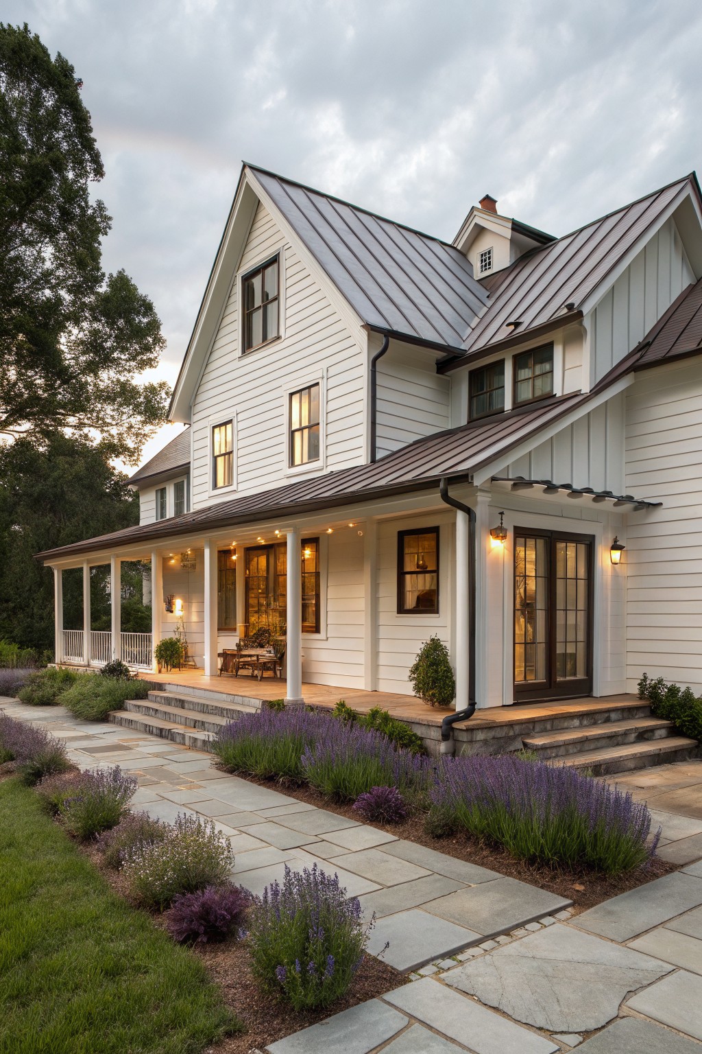 A two-story white farmhouse with a dark metal roof, covered porch supported by white columns, and a stone pathway bordered by lavender plants.