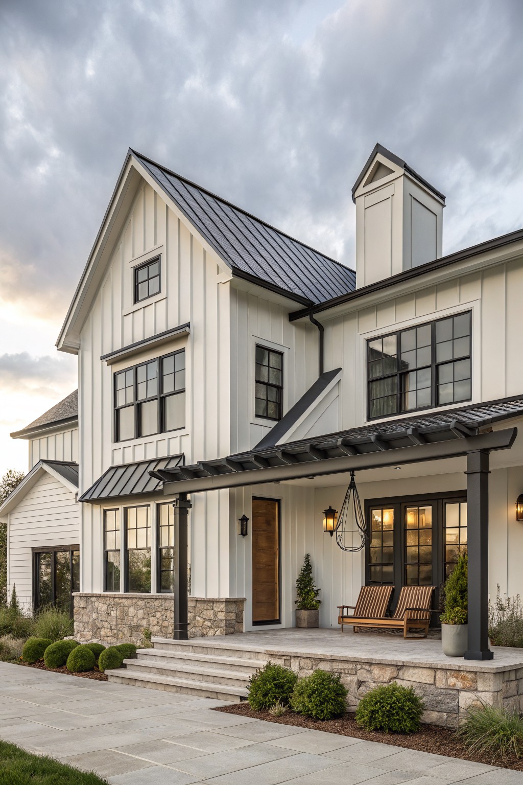White two-story farmhouse with a dark metal roof, black-framed windows, stone foundation, and covered porch with wooden seating.