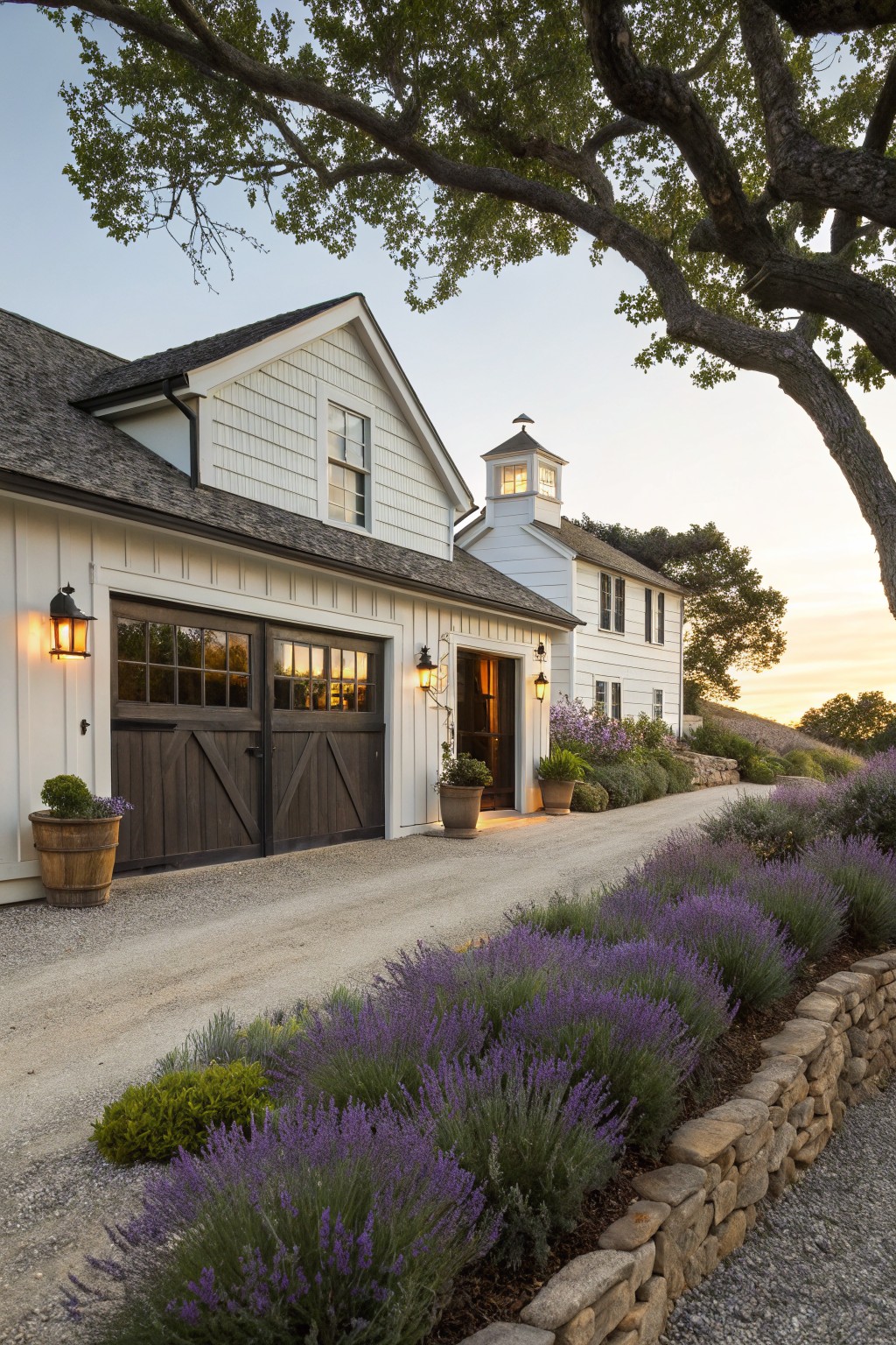 A white farmhouse exterior with dark barn-style garage doors, a gravel driveway bordered by purple flowering plants and stone edging, and a small cupola above the entry.