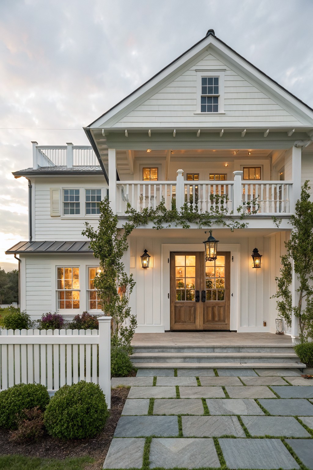 Front view of a white two-story farmhouse with a covered porch, double wooden entry doors, upper balcony, stone steps, and paved walkway.