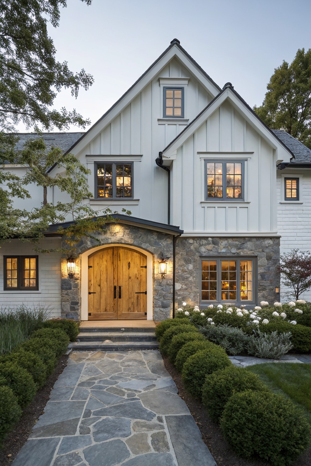 A white farmhouse exterior featuring a wooden double door set inside an arched stone surround, with lanterns on either side and windows above.