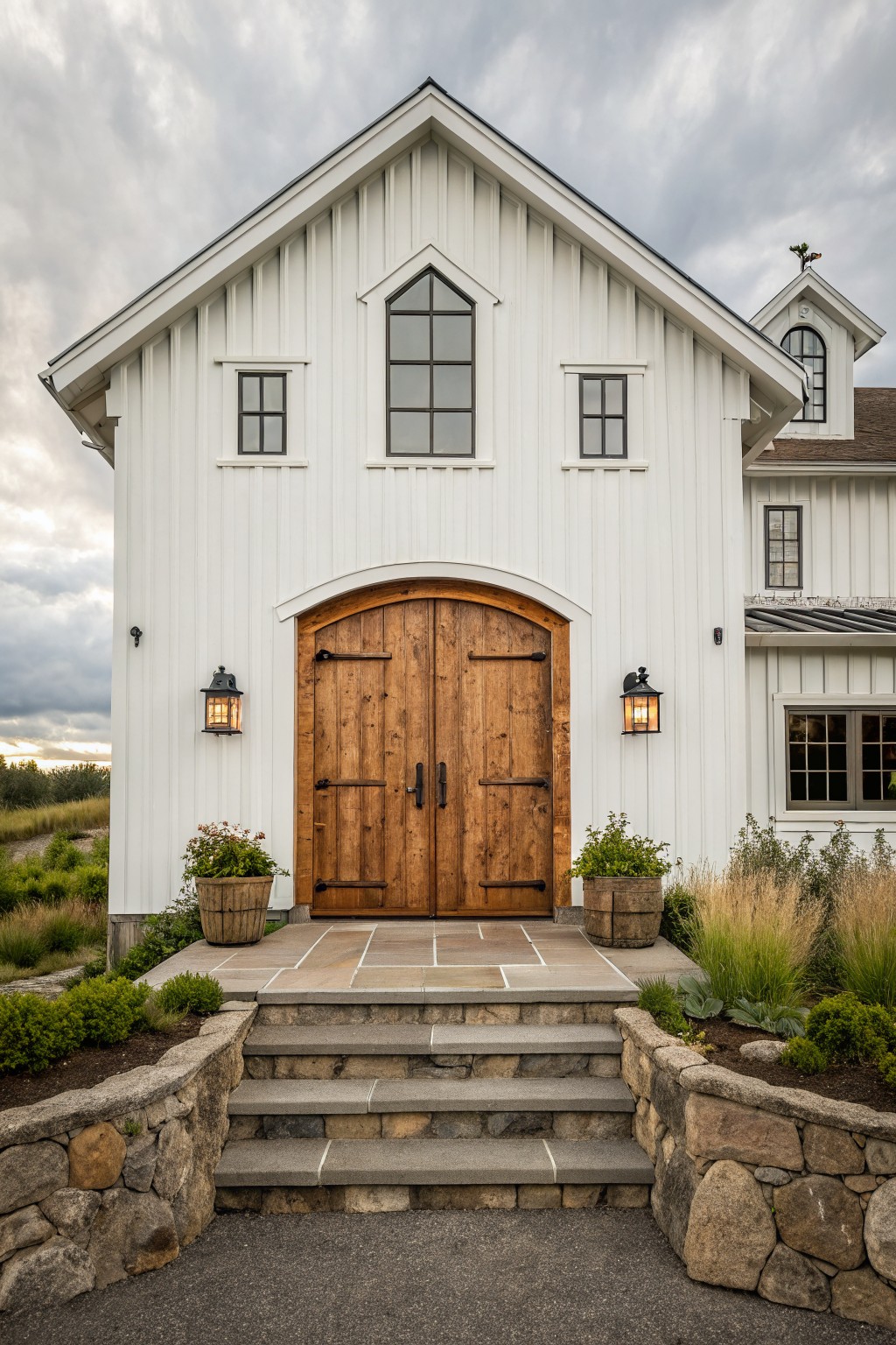 Front view of a white board and batten farmhouse with an arched wooden double door, stone steps, and two wall lanterns.