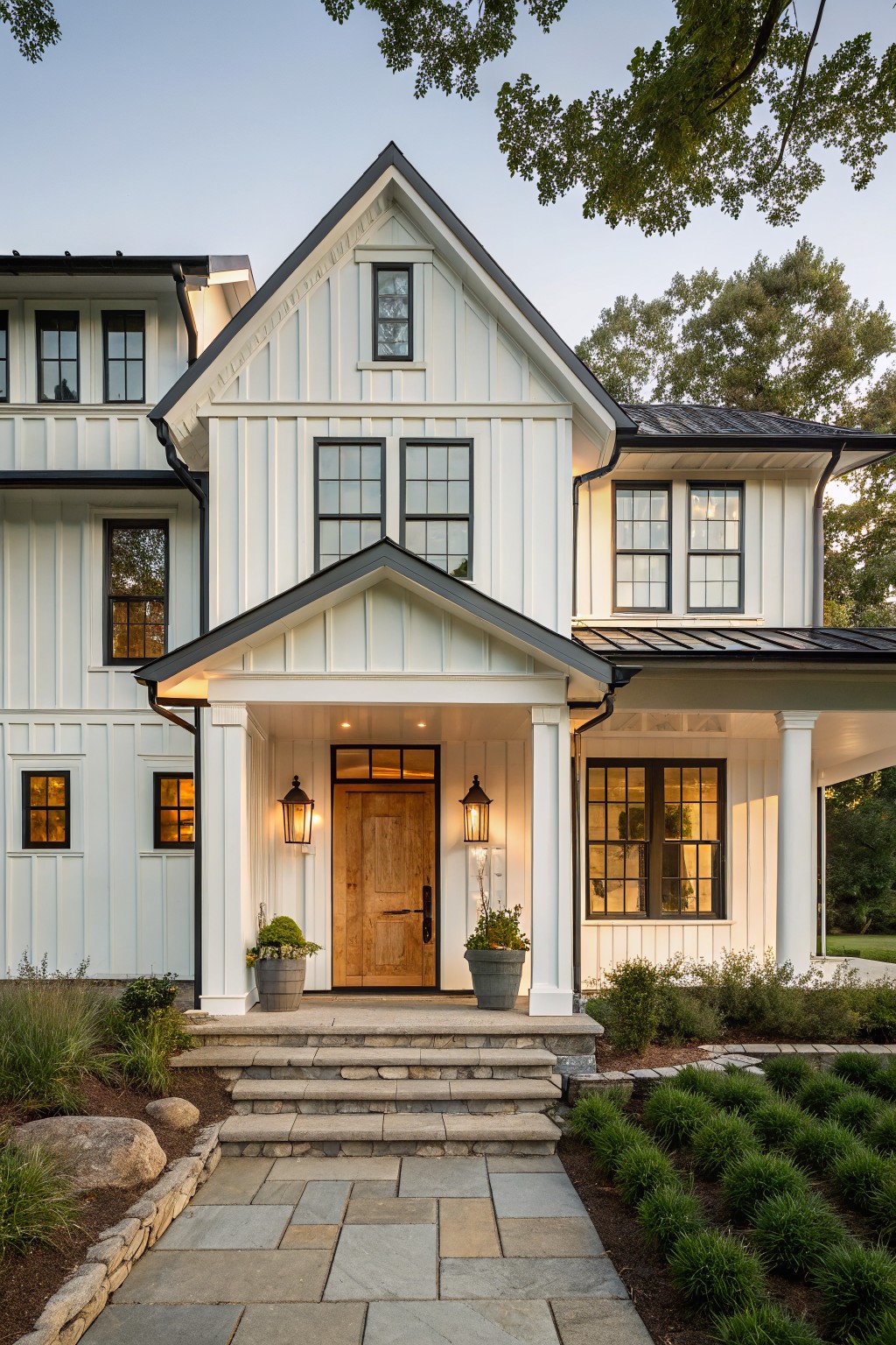 White two-story house with black trim, wooden door, stone steps, and evening lights