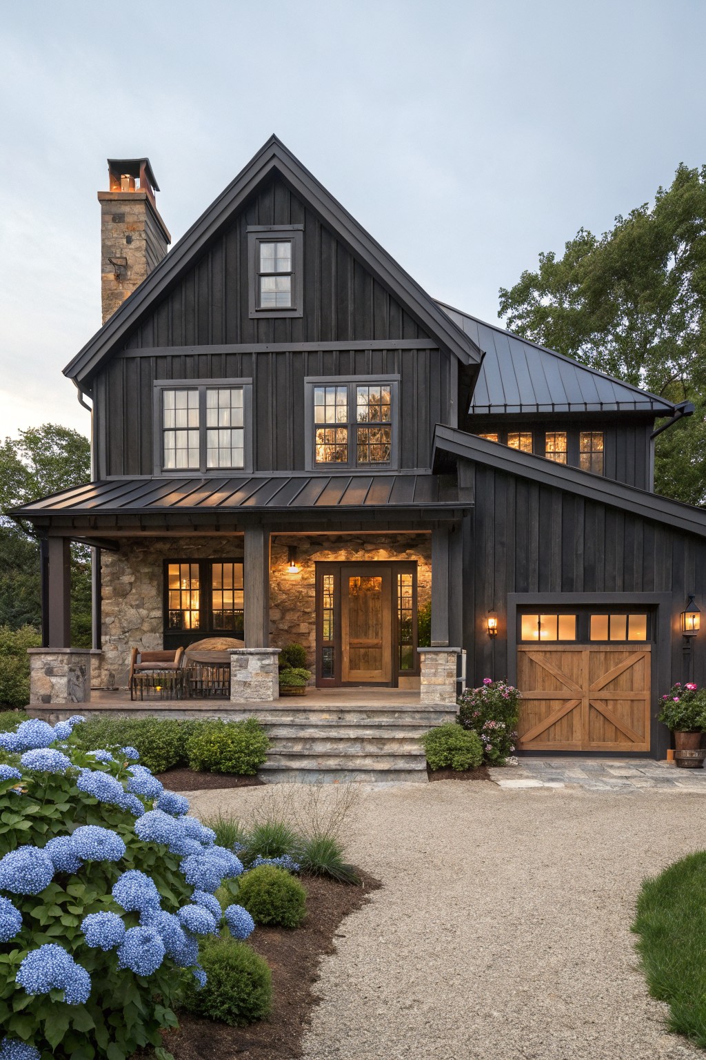 A two-story farmhouse exterior with dark board and batten siding, stone foundation, wooden garage door and front door, and blue hydrangeas along a gravel driveway.