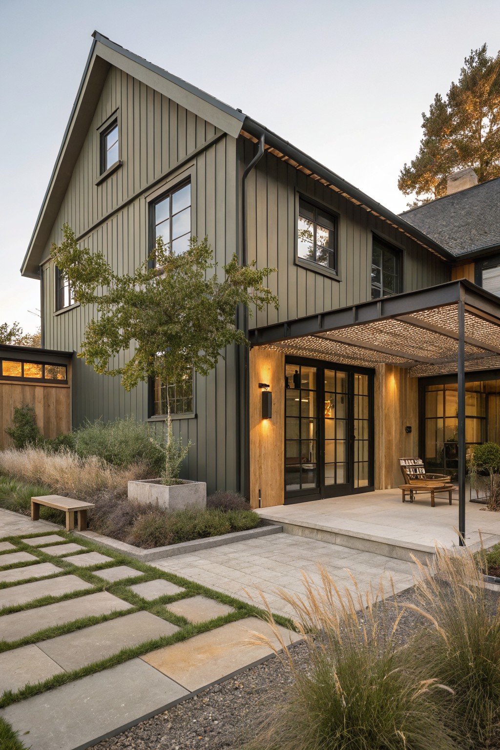 A two-story farmhouse exterior with dark green board and batten siding, black-framed windows, and a wooden entry door under a metal pergola.