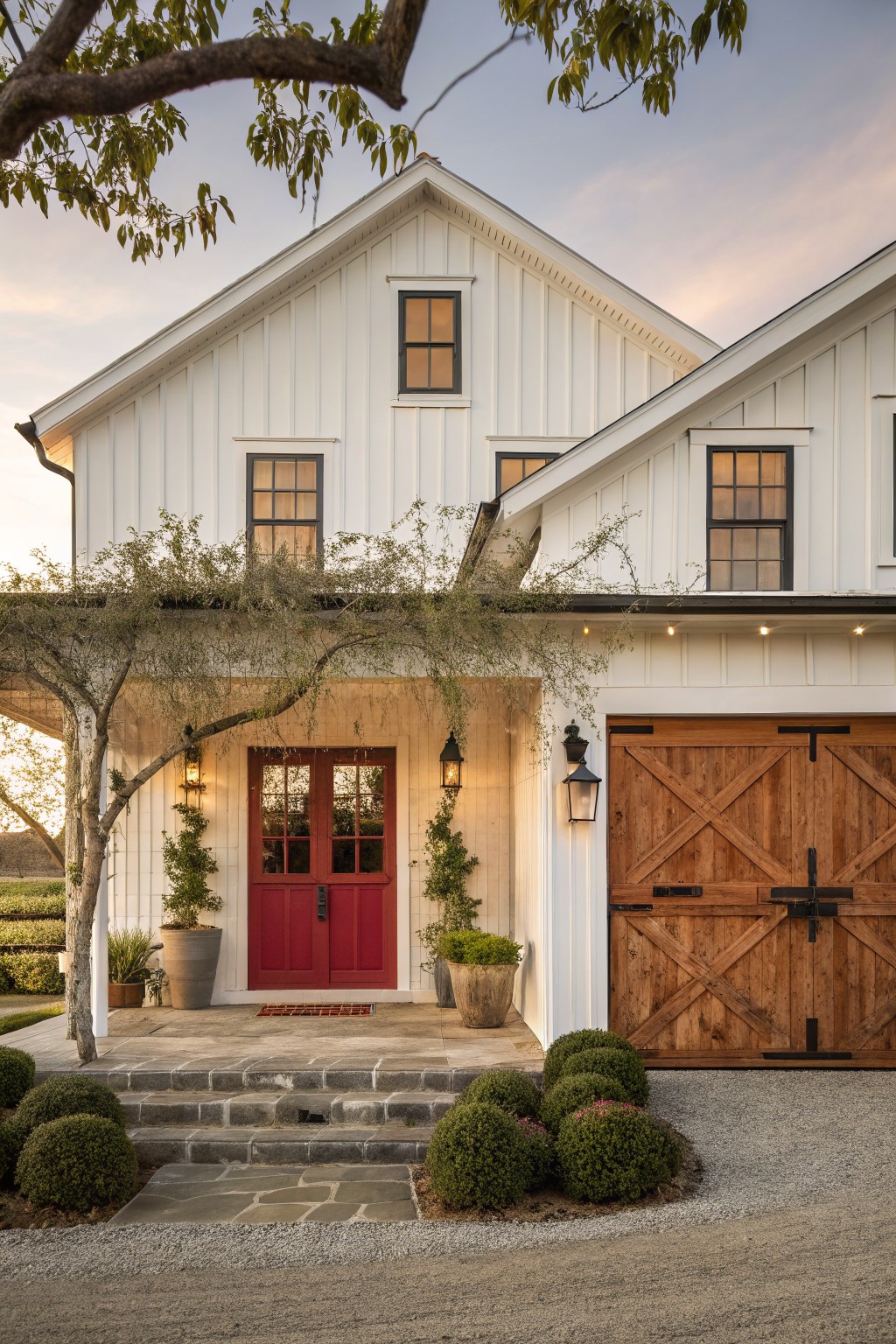 A white board and batten farmhouse exterior featuring a red front door, wooden garage doors, and stone steps.