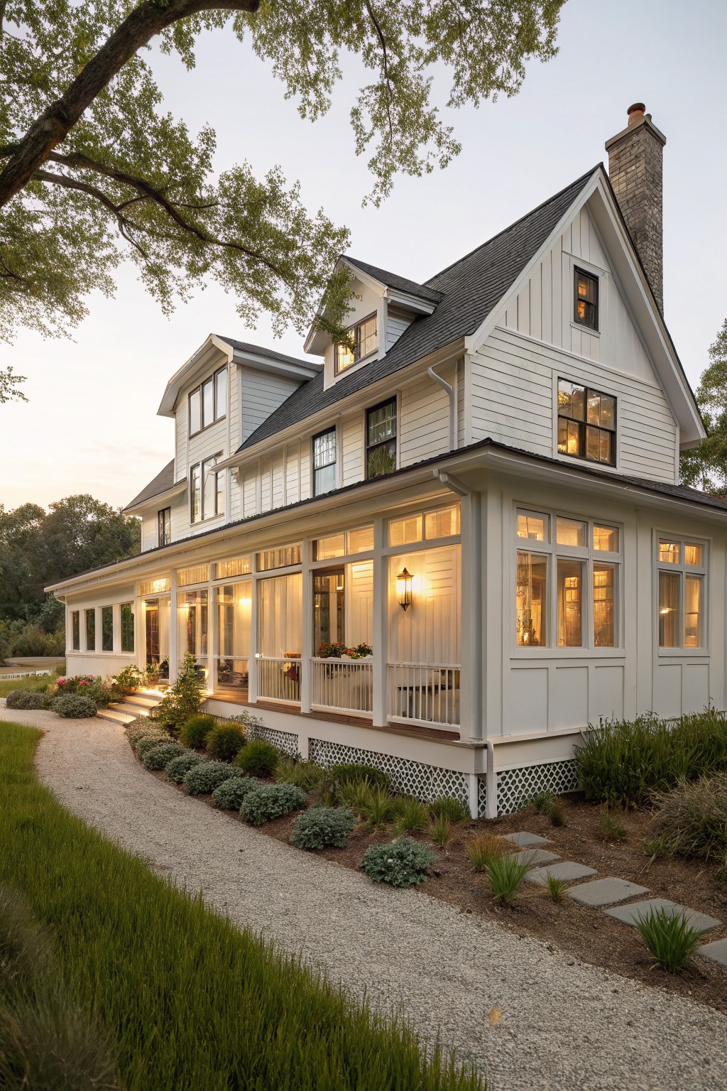 A two-story white farmhouse with board and batten siding on the gables, a wraparound porch with lights on, and a gravel path in front.
