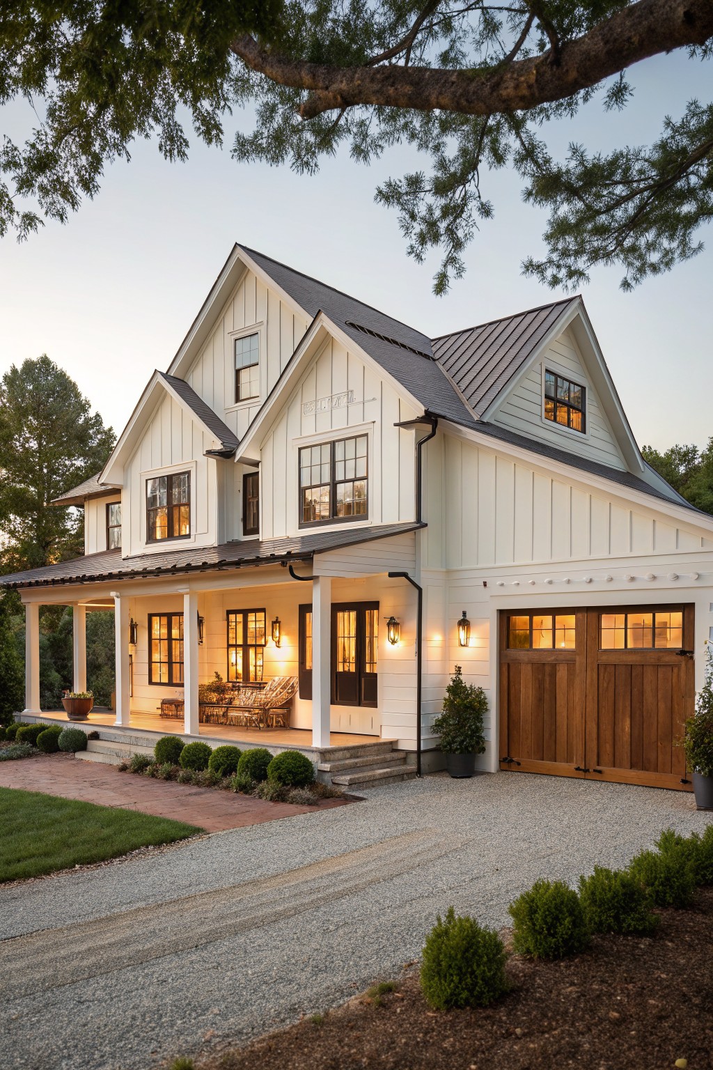 A white two-story farmhouse with board and batten siding, a covered porch, and brown wood garage doors at dusk.