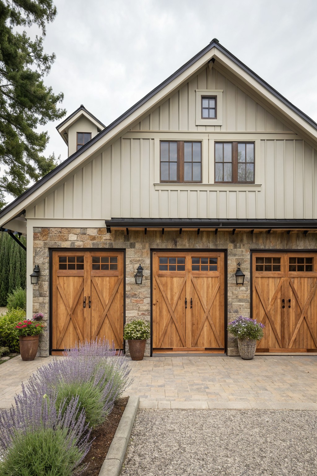 A farmhouse exterior featuring light board and batten siding on the upper level, a stone base, three wooden garage doors, and front landscaping with lavender and potted plants.