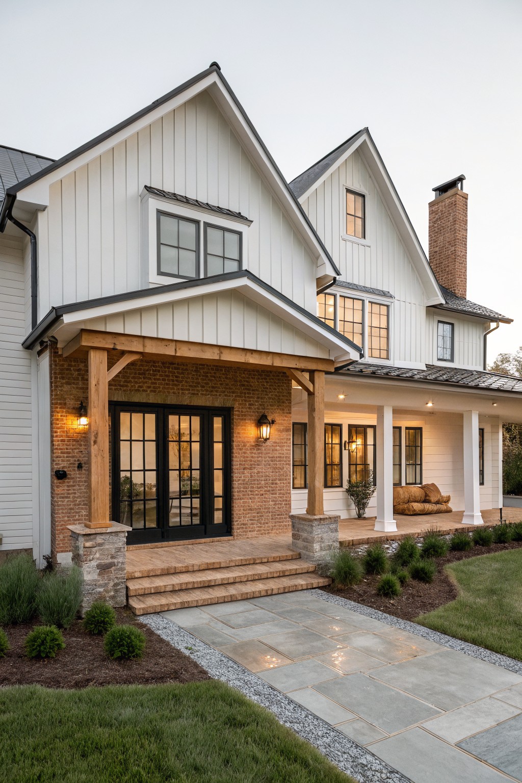 A two-story white farmhouse with board and batten siding, brick around the front entry with black doors, wooden porch beams, and a paved walkway in front.
