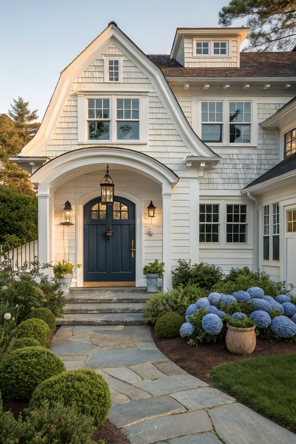 A white shingled house with a dark blue door centered under a white arched portico, stone steps, and a pathway bordered by blue flowering shrubs.