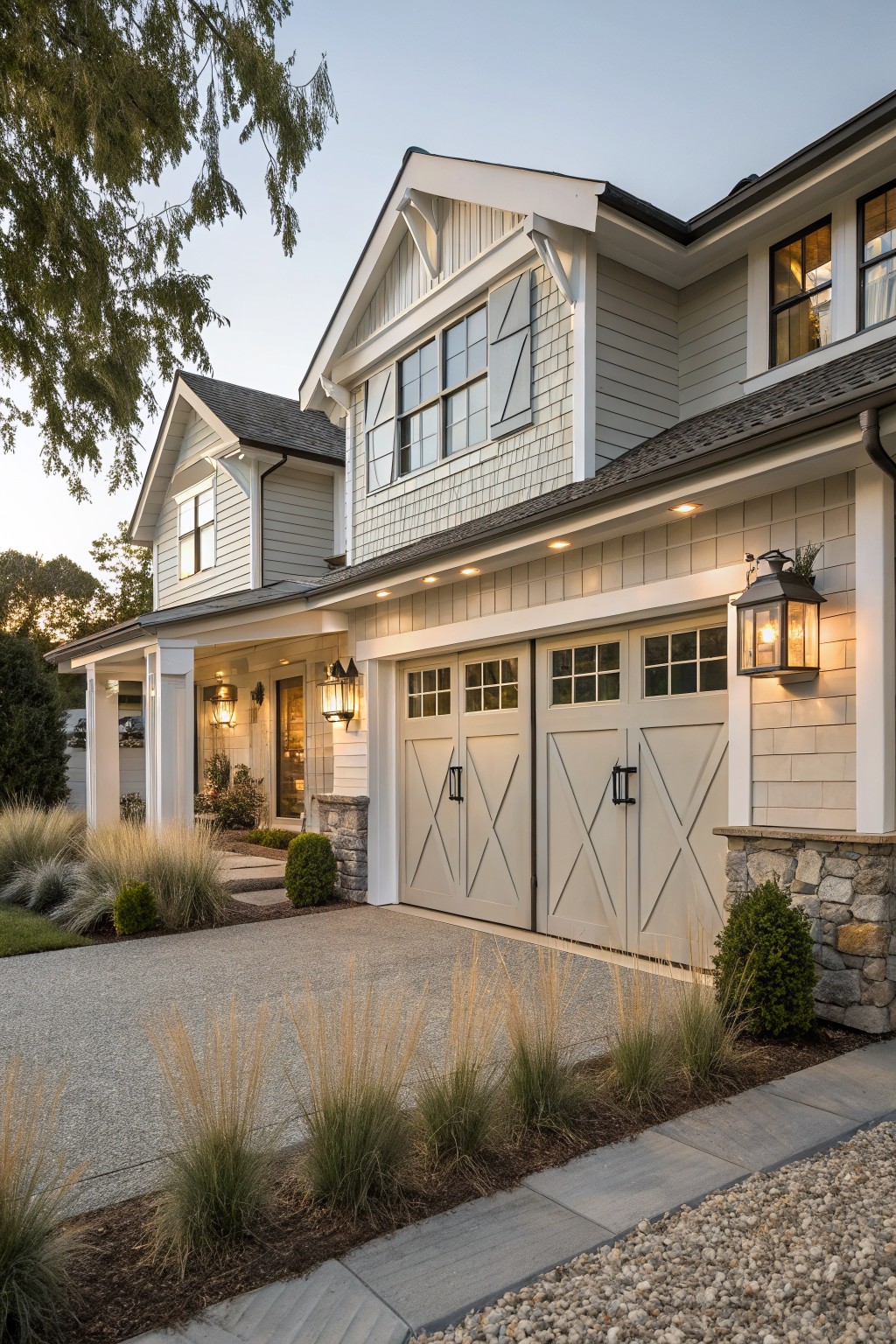 A two-story coastal farmhouse with light gray siding, white trim, stone veneer on the lower garage walls, and two X-pattern garage doors.