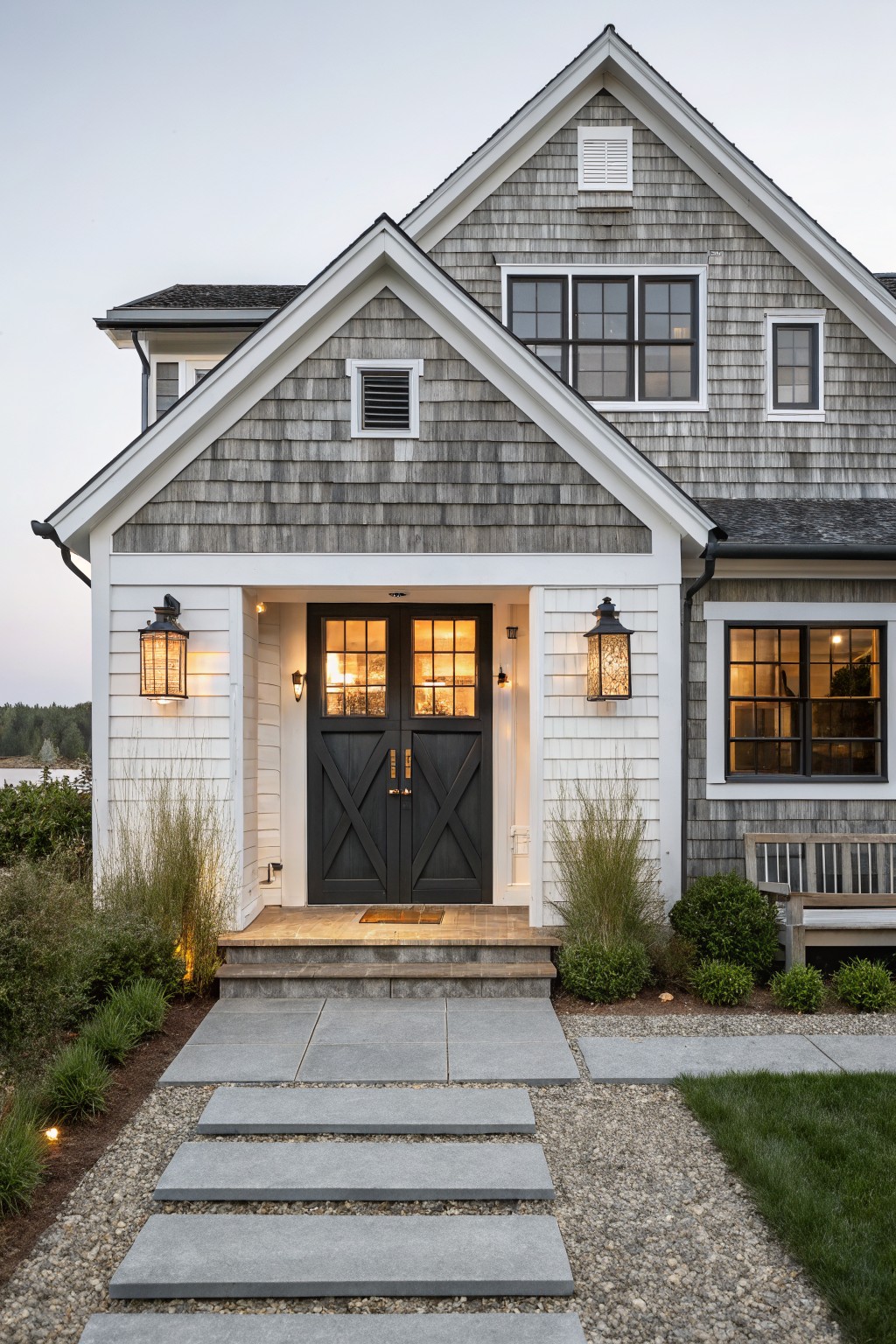 Front exterior of a coastal farmhouse showing gray cedar shingles, white trim, a dark double door entry, stone steps, and flanking lanterns.