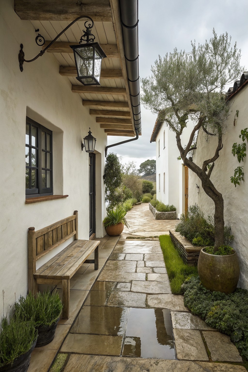Wet stone path in white-walled courtyard with wooden bench, lanterns, and potted plants.