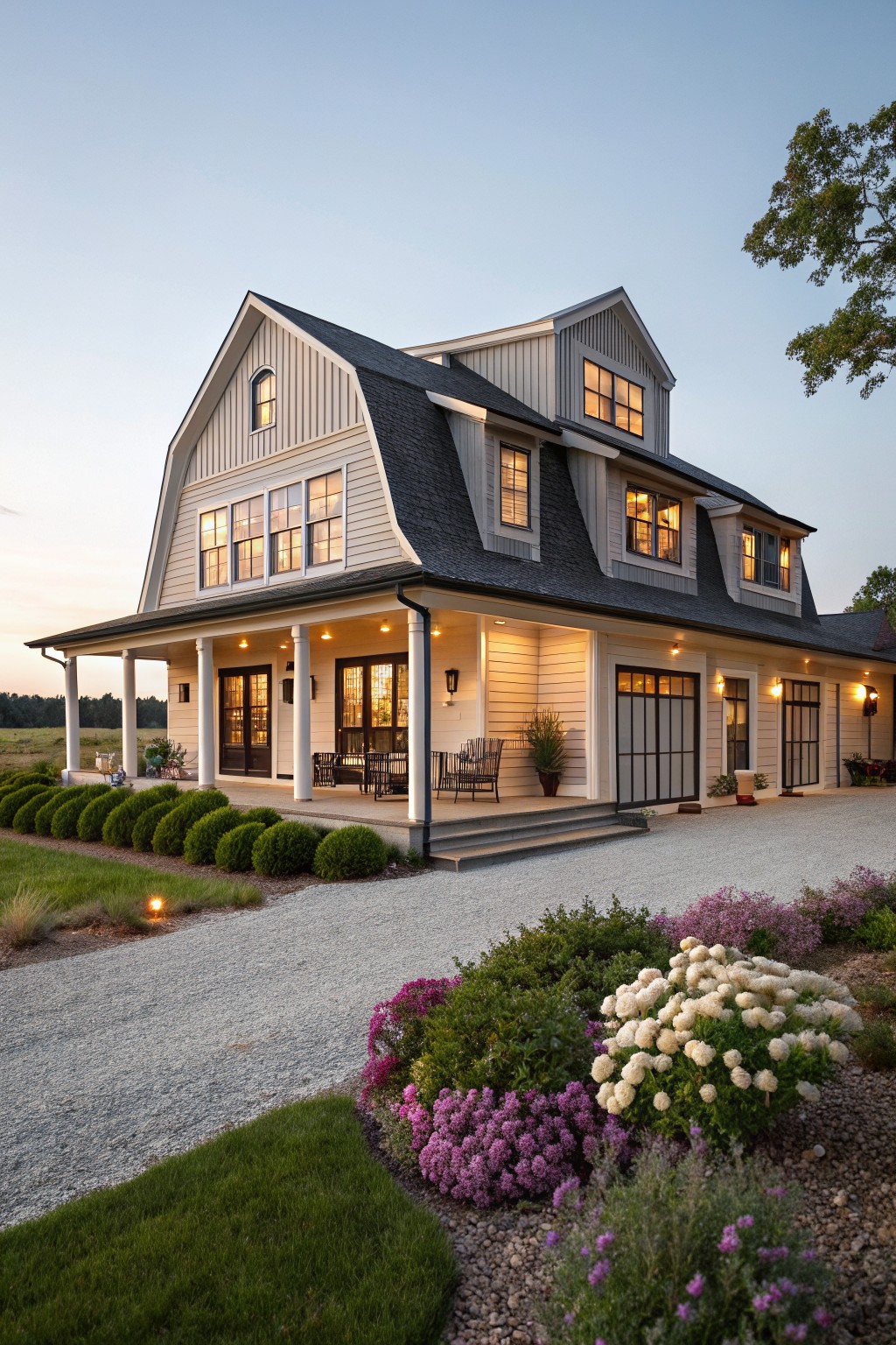 A two-story coastal farmhouse with light siding, a dark shingled roof, multiple gables, and a wide front porch supported by white columns, viewed from the gravel driveway at dusk.