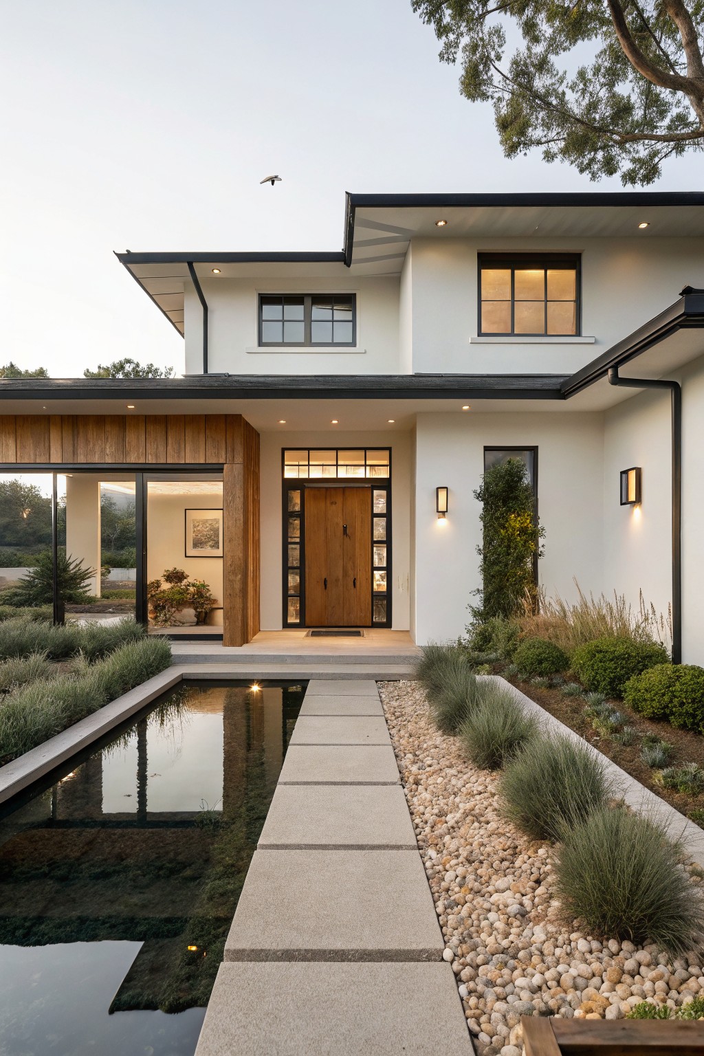 A modern white house with black trim and a wooden front door, approached by a concrete paver path that crosses a shallow reflecting pool with gravel and grasses on the right.