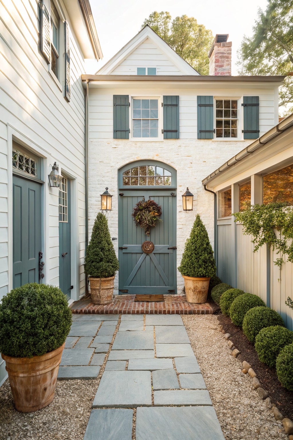 A front view of a white house with a central arched doorway, flanked by two potted evergreen shrubs on a stone pathway.